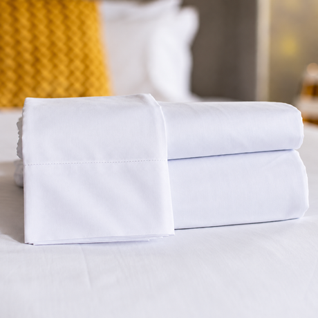 Pictured: Neatly folded white sheets on top of a white bed with a soft blur around the background. Two sheets are stacked while another is draped over the stack.