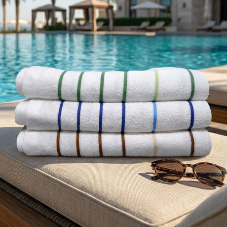 Pictured: Resort pool setting with three pool towels neatly folded in a stack on a chaise lounge with tortoise shell sunglasses folded in front of the stack. The colors of the towels from top to bottom are: Palm Green, Ocean Blue and Sand Beige.