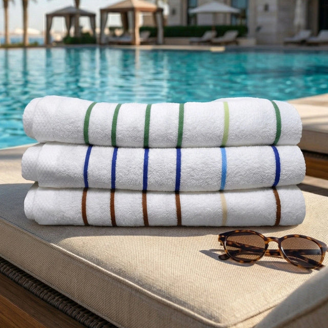 Pictured: Resort pool setting with three pool towels neatly folded in a stack on a chaise lounge with tortoise shell sunglasses folded in front of the stack. The colors of the towels from top to bottom are: Palm Green, Ocean Blue and Sand Beige.