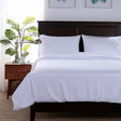 Pictured: Modern bedroom setting with a nightstand holding a plant, teacup, and silver lamp on the left. On the right is a dark wood bedframe with white bedding and pillows.