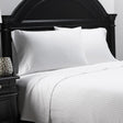 Pictured: Twill fabric white bedding on a neatly made bed with a black wood headboard. There is a matching nightstand to the left. The bedding is the Millennium T-250 Striped Sheets.