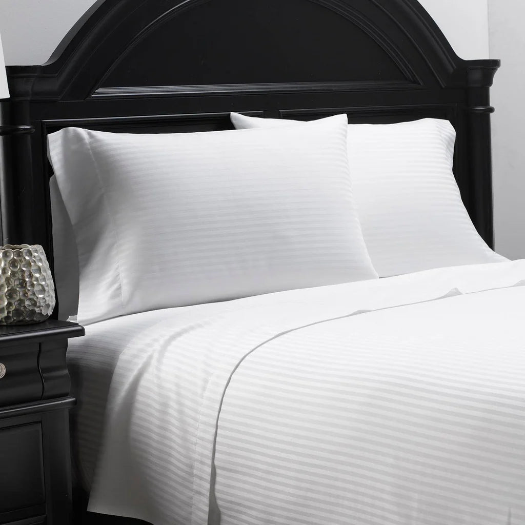 Pictured: Twill fabric white bedding on a neatly made bed with a black wood headboard. There is a matching nightstand to the left. The bedding is the Millennium T-250 Striped Sheets.