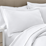 Pictured: Closeup of a pillow on a nicely made white bed against a beige headboard. The pillow is wearing he Millennium T-250 Pillow sham in the Stripe style. It  is made with a twill fabric to create the pattern.