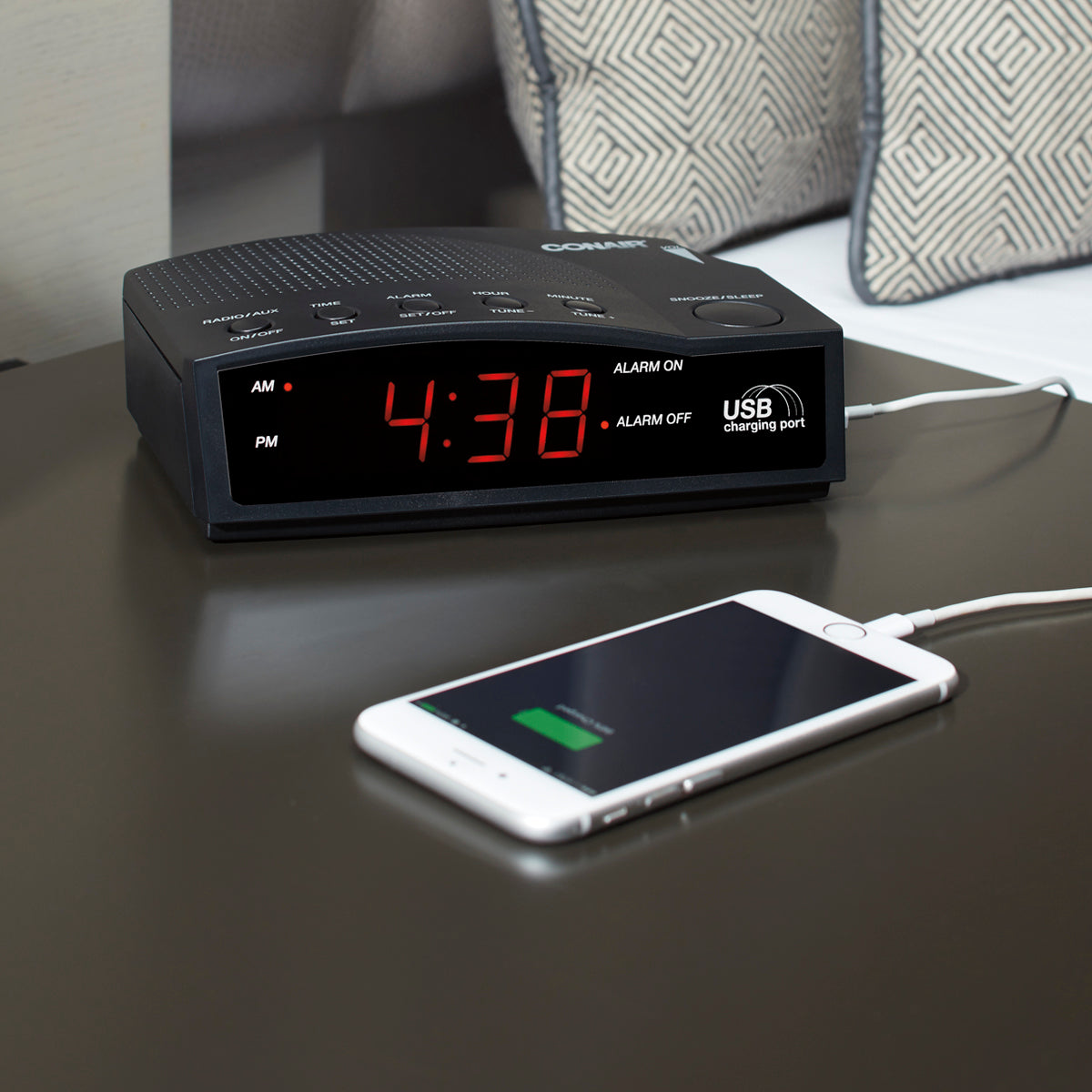Black alarm clock with digital display on a surface, next to a smartphone charging cable.